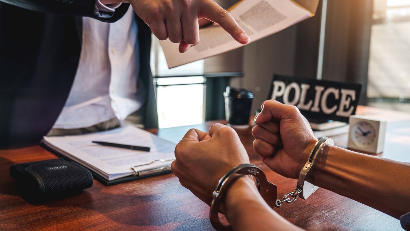 A person in handcuffs sitting at a desk while another individual points at them, indicating legal or police action in a formal setting.