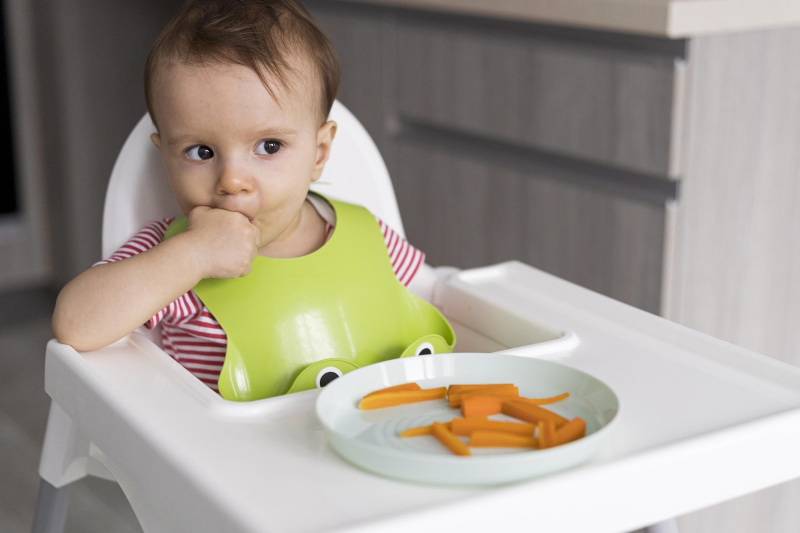 A baby eating sliced carrots on her high chair