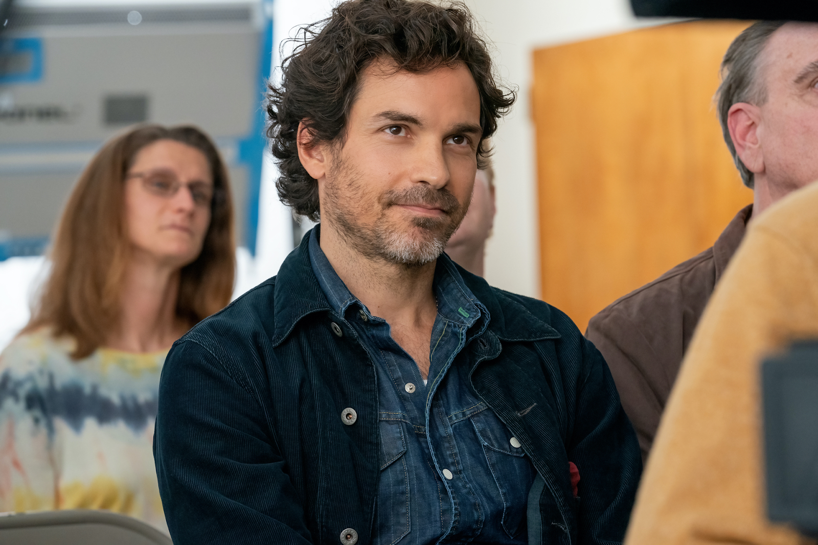 Santiago Cabrera en una escena de una serie, con expresión seria, vistiendo una chaqueta de mezclilla sobre una camisa casual.