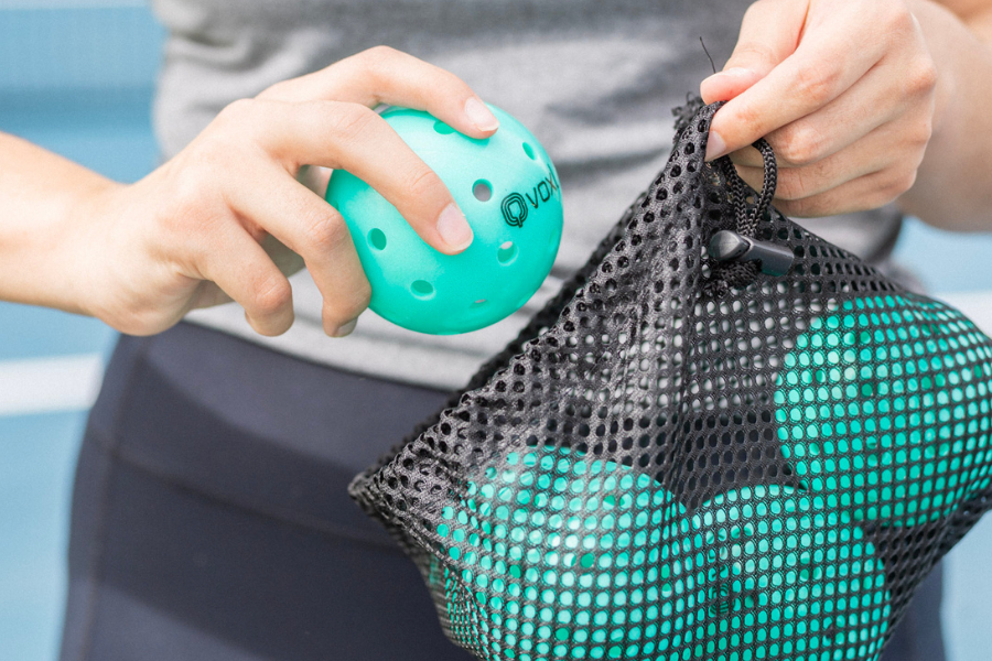Close-up of a player pulling a green Voxl pickleball out of a black mesh gear bag on a court.