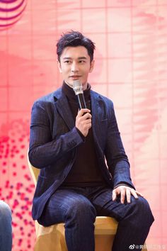 This contains an image of Huang Xiaoming sitting on a chair holding a microphone in his hand and speaking into the microphone