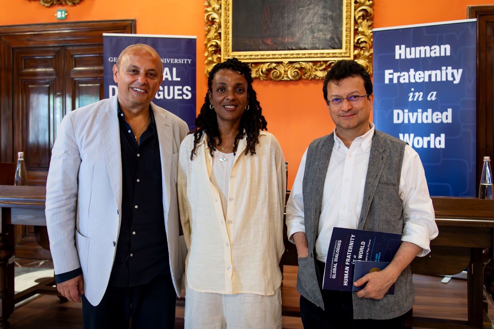 Dean Masri stands next to authors Nasrine Malik and Ranjit Hoskote after their panel discussion, in front of Global Dialogue conference branding and a large gold framed painting.