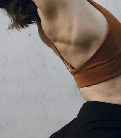 A close-up of a person’s torso, wearing an orange top and black bottoms. The focus is on the side profile of the torso, highlighting the well-defined ribcage and abdominal muscles.