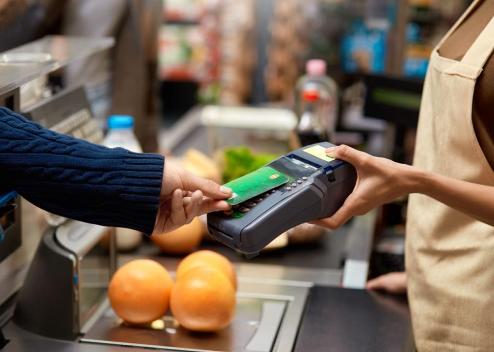 A close-up of a transaction at a grocery store checkout, where a customer is making a payment using a credit card, tapping it on a card reader held by a cashier. 