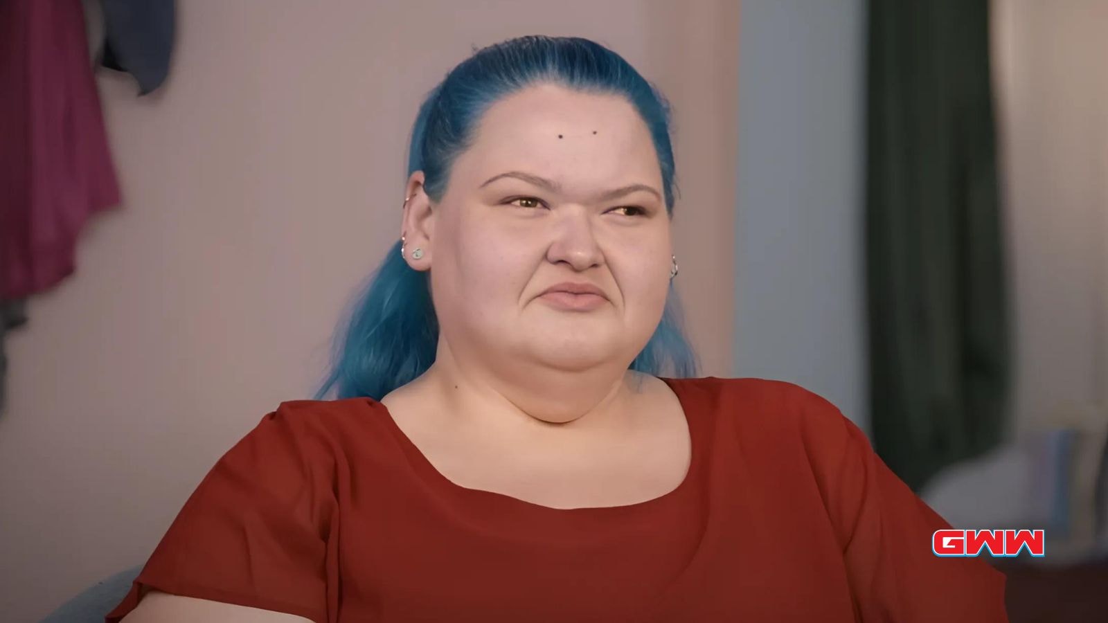 Amy with blue hair wearing a red shirt, appearing thoughtful while sitting indoors.