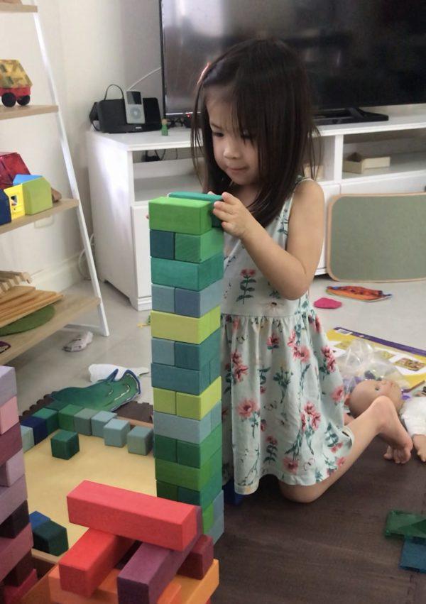 A child carefully choosing single-size blocks from Grimm’s Large Stepped Pyramid set and stacking them to form a tall tower with cool-tone green blocks only