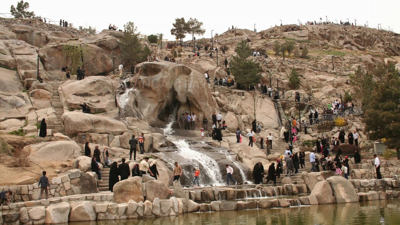 Busy scene at Koohsangi Park in Mashhad, Iran, showcasing a rocky waterfall, stairways, and visitors exploring the natural landscape, surrounded by lush greenery and rocky hills.