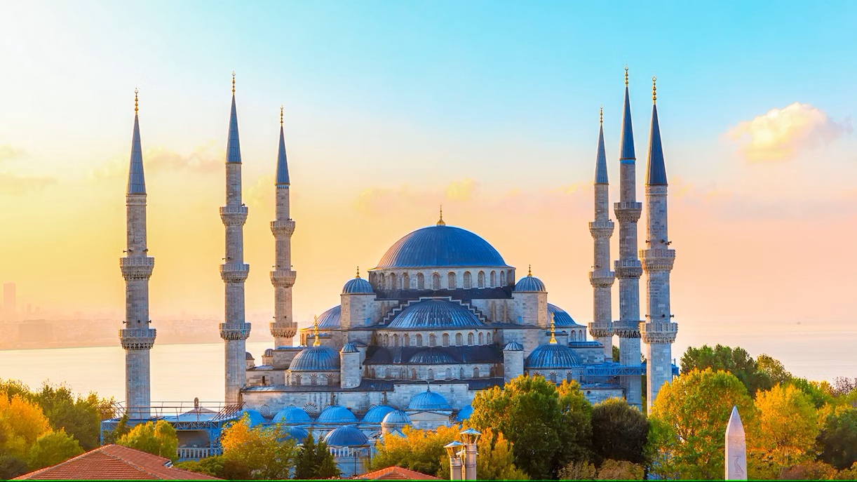 Scenic view of the Blue Mosque (Sultan Ahmed Mosque) in Istanbul, Turkey, at sunset with six minarets, domes, colorful sky, and lush green trees.