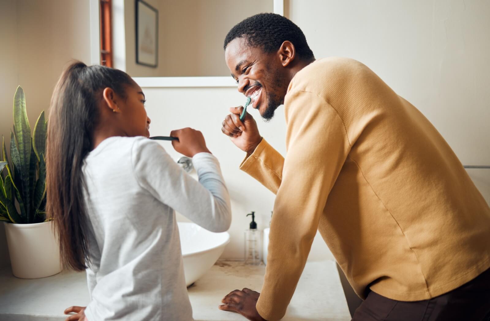 A father and his school-aged daughter looking at each other and smiling while brushing their teeth over the sink in the bathroom