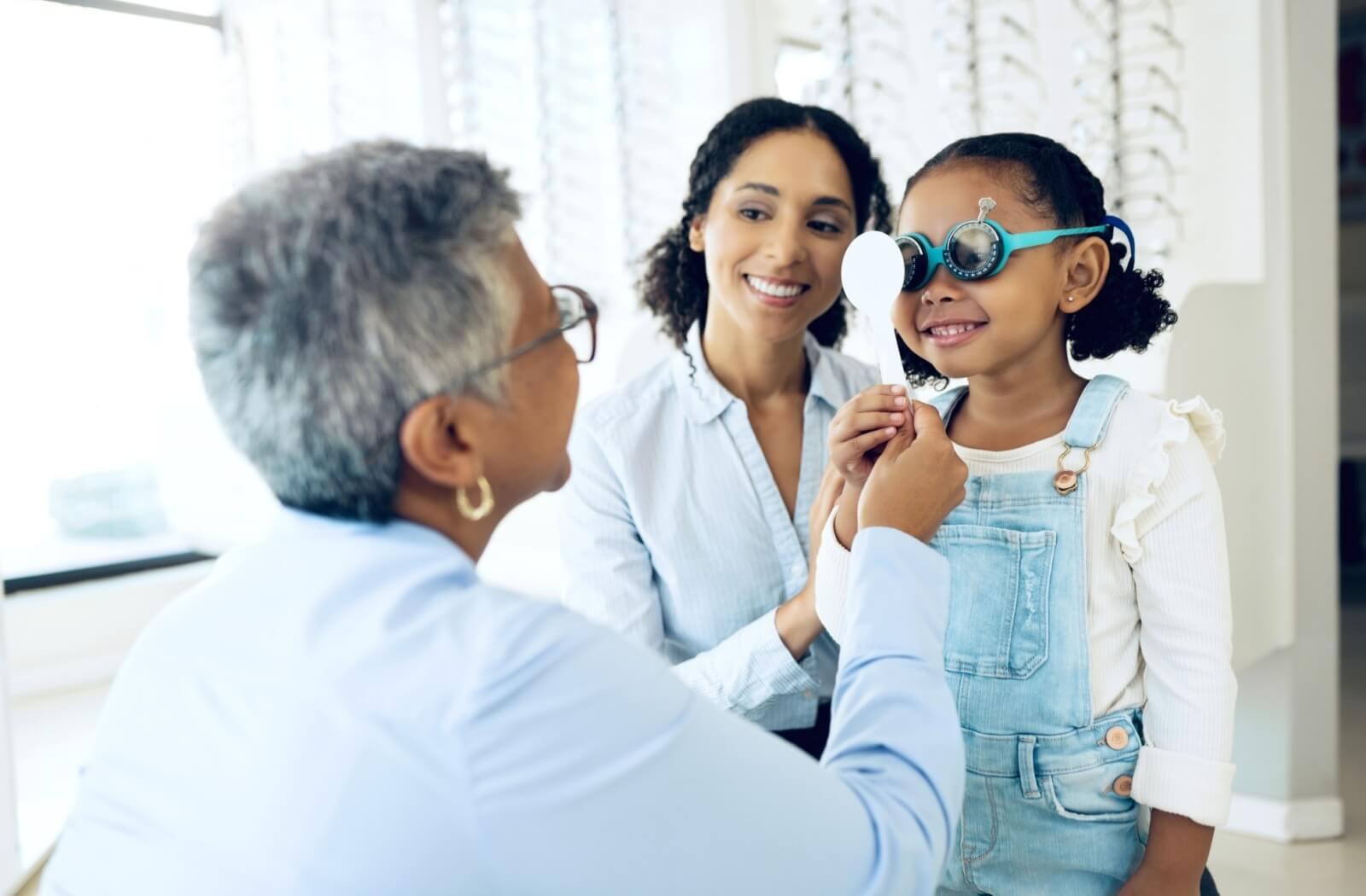 An optometrist covers one of their child patient's eyes during an exam while their mother sits nearby.