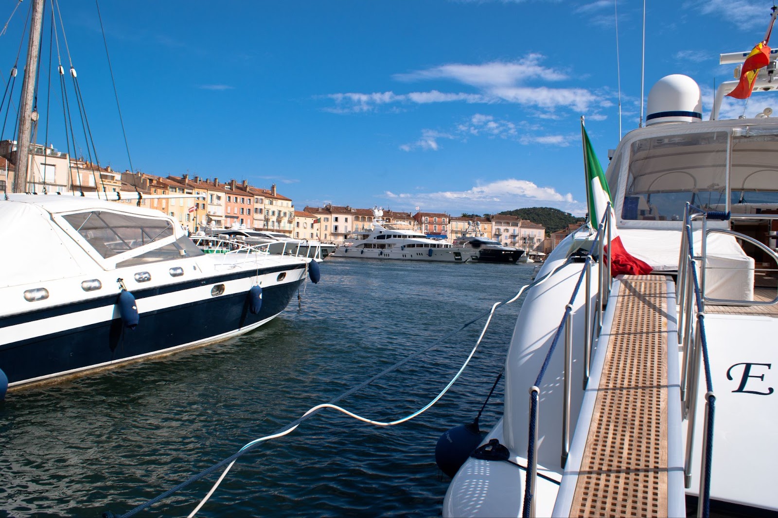 Docked luxury yachts in the harbor of Saint-Tropez with Italian flag, colorful waterfront buildings, and additional yachts in the background under a sunny blue sky.