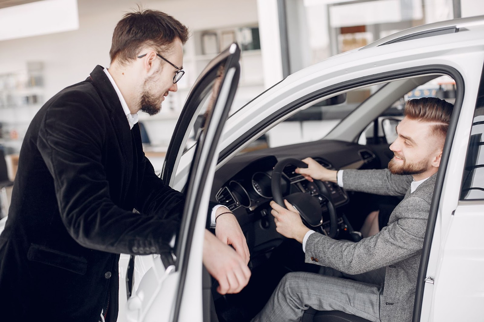 Two men in a car dealership, one sitting inside the car discussing features with the other standing outside