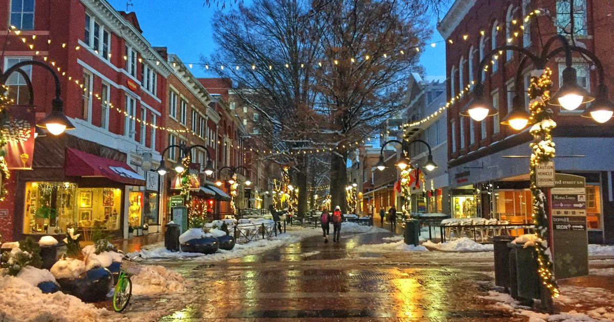 A view of a Charlottesville street, snow visible on the ground and shoppers walking in the street. Lights are strung up from light poles.