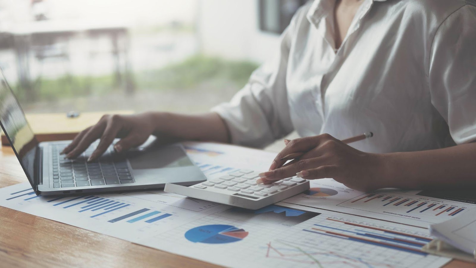 Close-up view of a professional working at a desk with a laptop and calculator, analysing business metrics and financial charts, highlighting the role of a skilled bookkeeper in managing business finances.