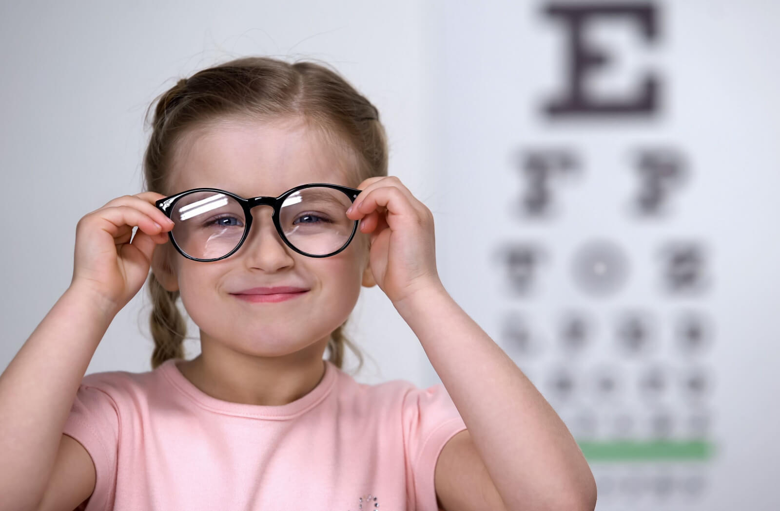 A little girl standing in front of an eye chart smiling while trying on a pair of myopia control glasses.
