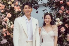 This  contain Hyun Bin and Son Ye Jin  standing next to each other in front of a flower covered wall