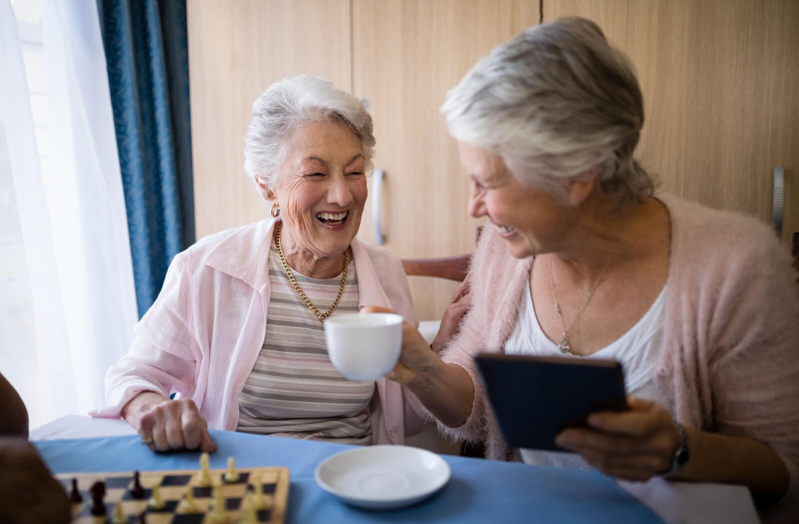 2 senior women drinking coffee and playing a friendly game of chess.