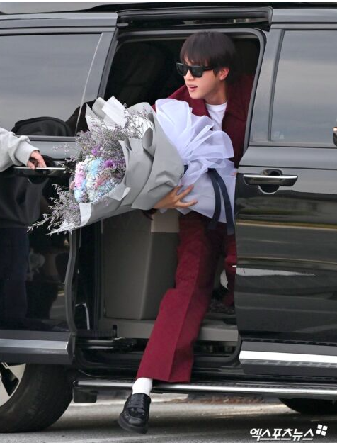 This contains an image of Jin coming out of a car, holding a flower wearing a burgundy suit and statement sunglasses