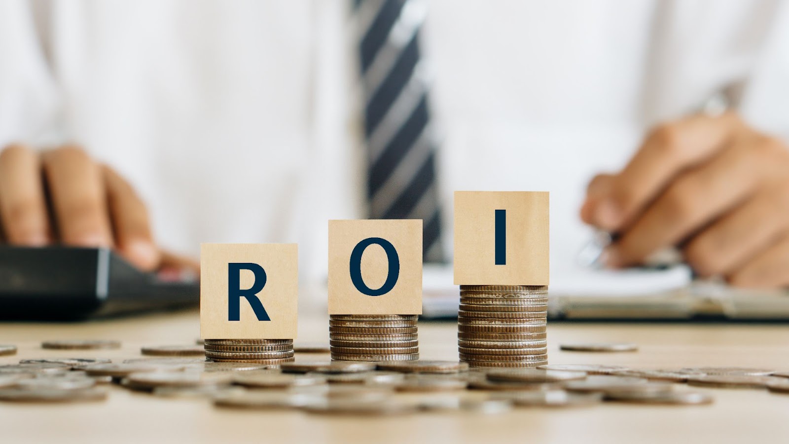 The word “ROI” is written on wooden cubes stacked on piles of coins with a man in a tie in the background. 