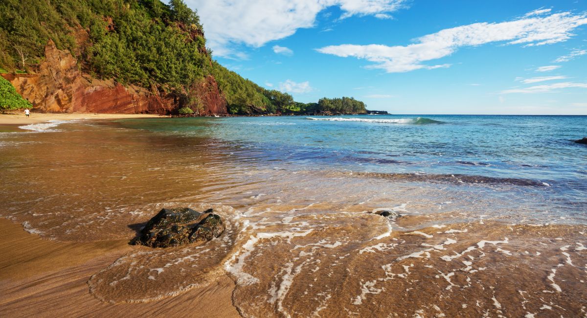 Red sand beach on a clear sunny day with clear turquoise waters, gentle waves, and a scenic backdrop of lush, red cliffs partially covered with green foliage, under a bright blue sky with sparse clouds.