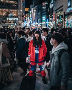 This contains a woman standing in the middle of a crowded city street at night wearing an orange jacket 