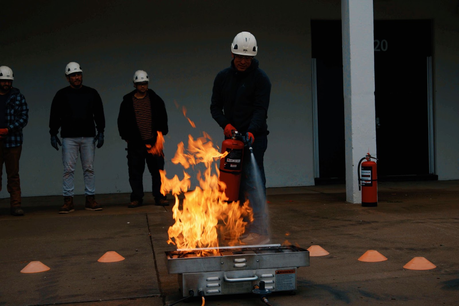 An individual in a safety helmet and gloves using a fire extinguisher to put out a controlled fire during a fire safety training exercise. A group of onlookers, also in helmets, stands in the background, observing the demonstration in an outdoor setting.