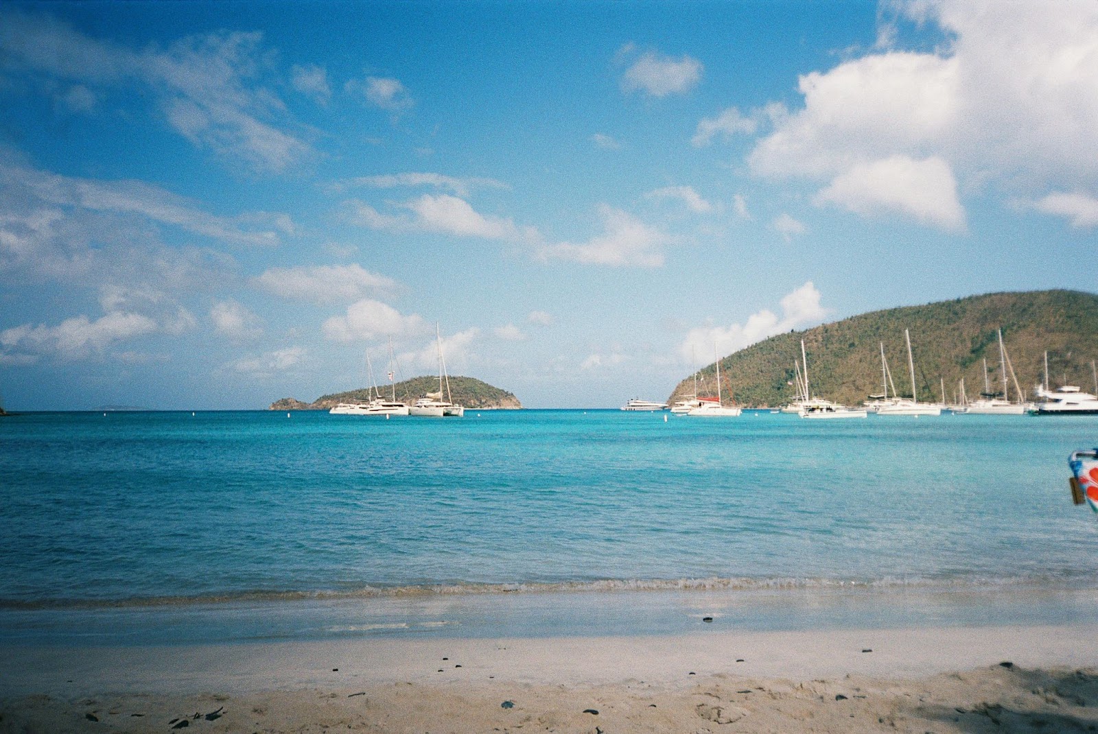 Yachts anchored near a sandy beach with calm turquoise waters and a hilly island in the background — typical of the British Virgin Islands sailing experience.