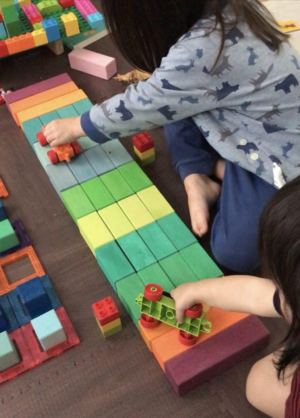 Two toddlers are engaging in a pretend play session with LEGO trucks and wooden blocks. The wooden blocks are lined up to make a 4 unit ramp. 
