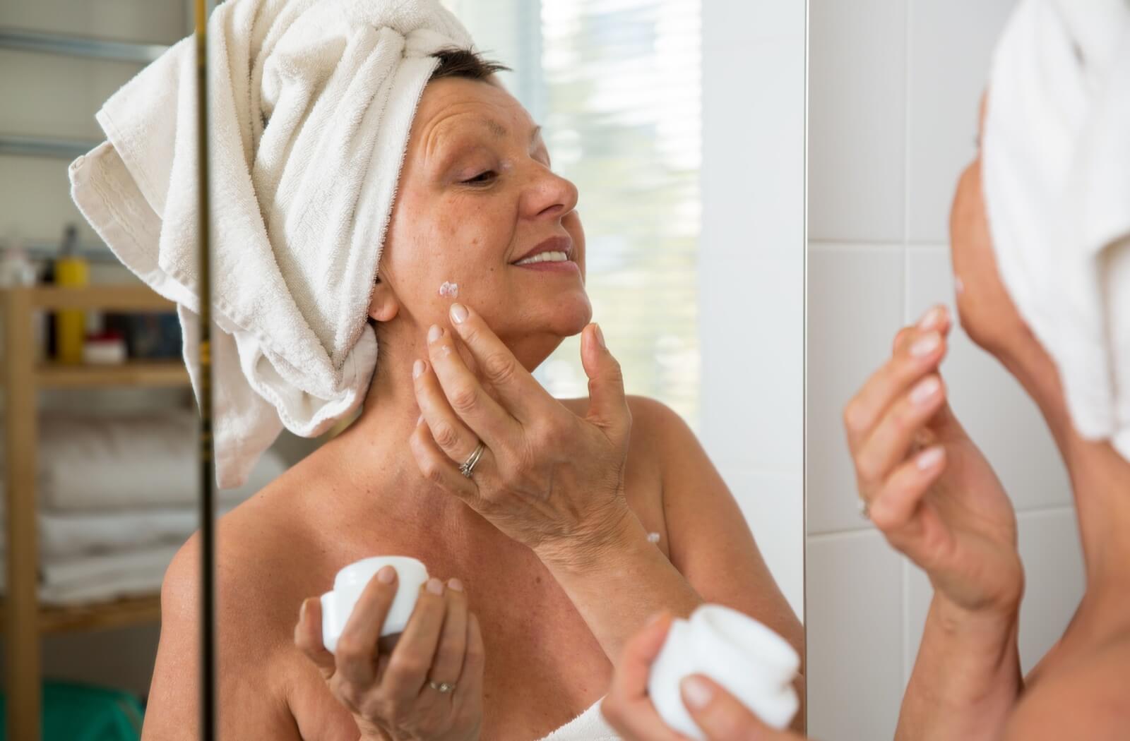 An older adult applies cream to their face while their skin is still soft following a shower.