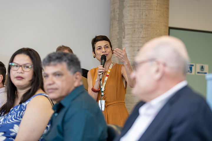  Nesta imagem, uma mulher com vestido laranja e crachá está em foco, falando ao microfone e gesticulando com uma das mãos. Ela parece estar conduzindo uma apresentação, palestra ou discurso, demonstrando confiança e engajamento.  Na parte frontal da imagem, há algumas pessoas parcialmente visíveis, sentadas e ouvindo atentamente. Um homem de óculos e blazer azul está desfocado no primeiro plano, o que dá destaque à oradora. Outra pessoa com roupas coloridas (azul e branco) também aparece na lateral esquerda.  O ambiente continua semelhante às imagens anteriores, sugerindo que todas fazem parte do mesmo evento em um local interno, com uma atmosfera formal e profissional.