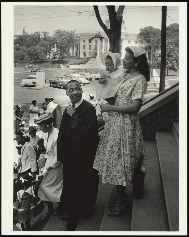 Image: Black-and-white photograph of Rev. Martin Luther King Jr. and his wife Coretta Scott King holding their infant daughter Yolanda Denise King while they stand on the steps in front of Dexter Avenue Baptist Church in Montgomery, AL. In the background are congregation members of the church where King pastored. Photograph by Moneta Sleet Jr., Johnson Publishing Company Archive. Courtesy J. Paul Getty Trust and Smithsonian National Museum of African American History and Culture.