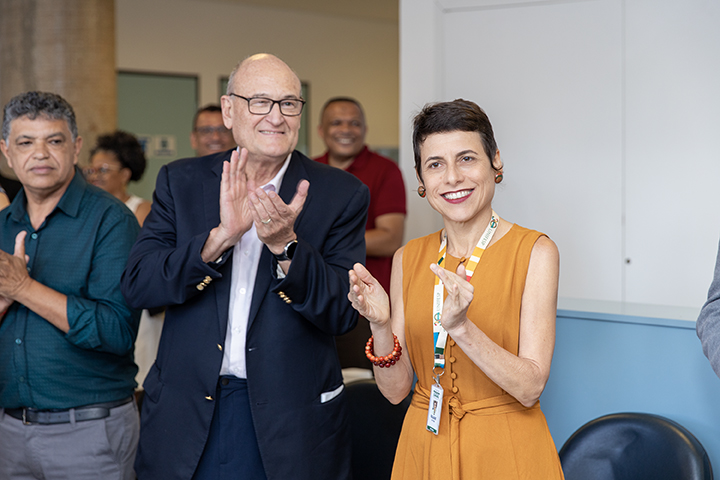 Esta imagem mostra um grupo de pessoas em pé, participando de um evento ou comemoração. Em primeiro plano, vemos um homem idoso, careca e de óculos, vestindo um blazer azul escuro e camisa branca. Ele está sorrindo e aplaudindo. Ao seu lado, uma mulher usando um vestido laranja, colar vermelho e crachá também sorri e aplaude, transmitindo entusiasmo e alegria.  Ao fundo, outras pessoas podem ser vistas sorrindo e participando do evento, sugerindo um clima positivo e descontraído. O ambiente parece ser o mesmo da imagem anterior, com paredes brancas e um espaço organizado e iluminado.