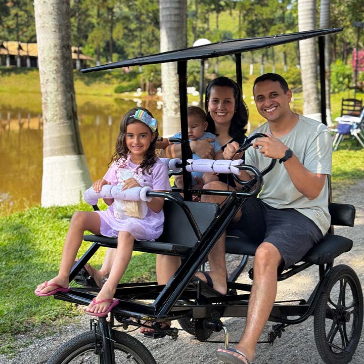Família feliz passeando de triciclo no parque com as crianças e pai sorrindo ao lado, cenário natural com árvores e lago ao fundo.