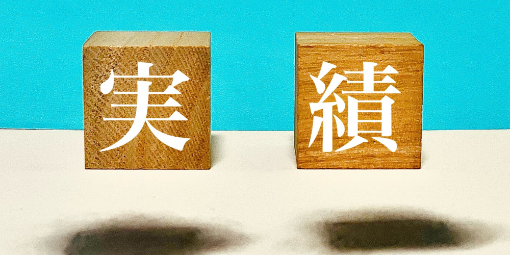  水色の背景と白い机の上に置かれた木製キューブ２個に、白文字で「実」「績」が表示されているシンプルな写真