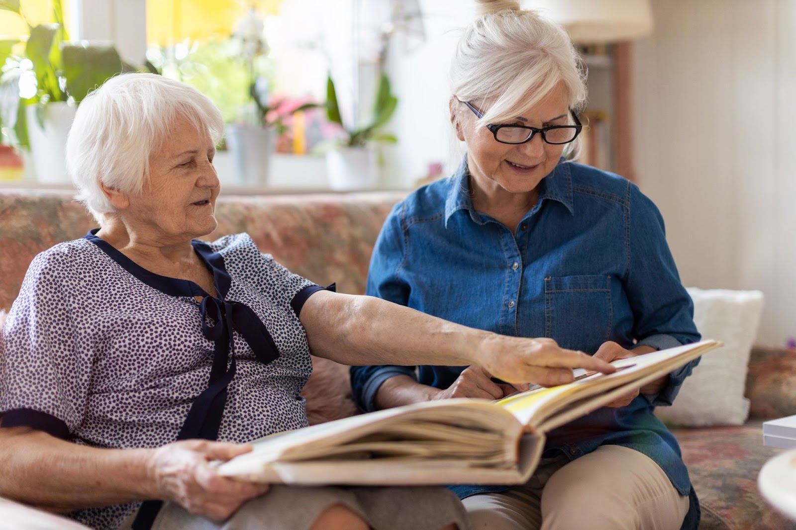 Senior woman and her adult daughter looking at photo album together on couch in living room.