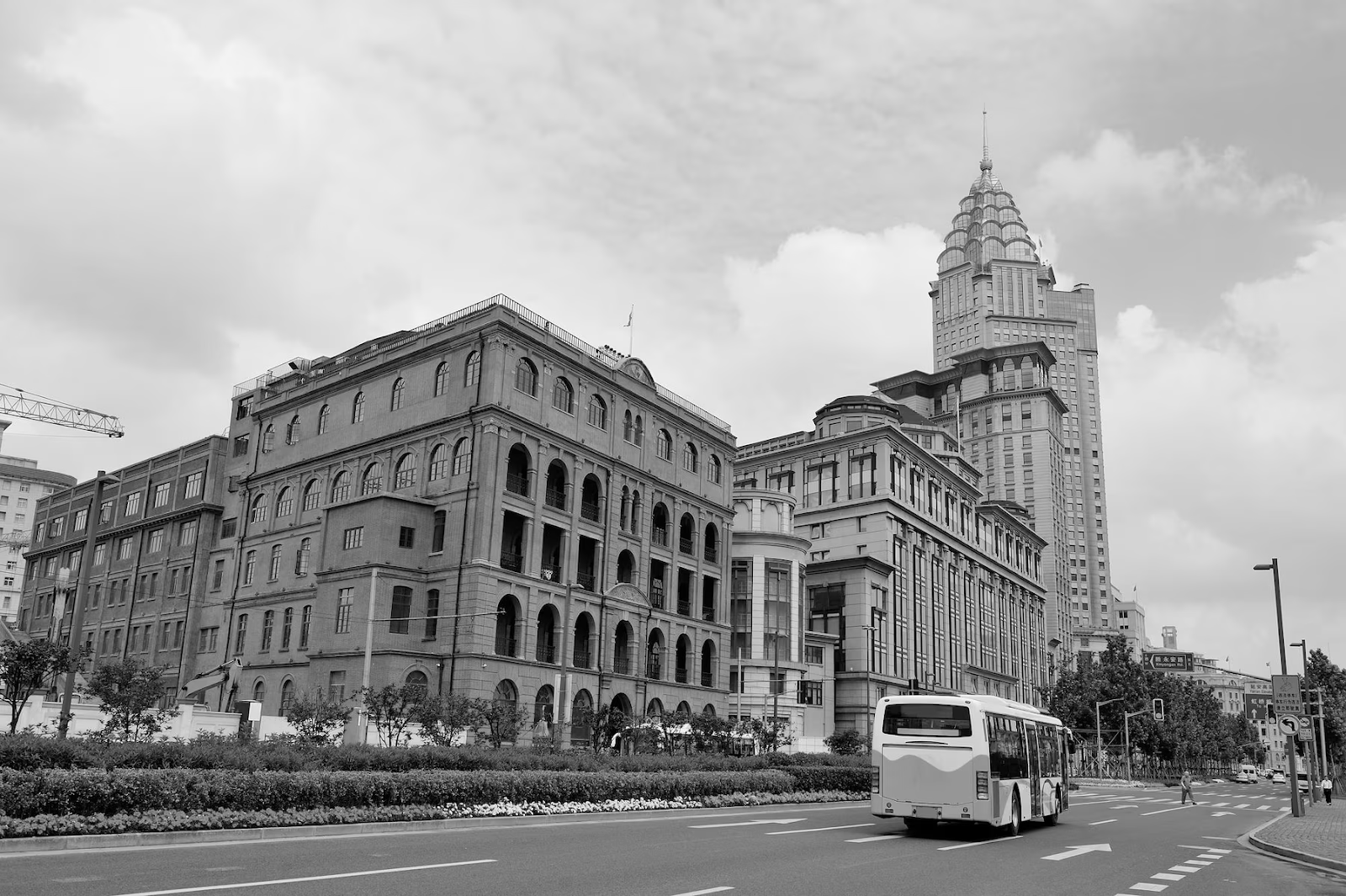 A black and white photo captures an urban street with a historic brick building juxtaposed against a tall, ornate skyscraper. A bus glides along the road under the partly cloudy sky, evoking the essence of architectural journalism as if documenting the city's evolving skyline.