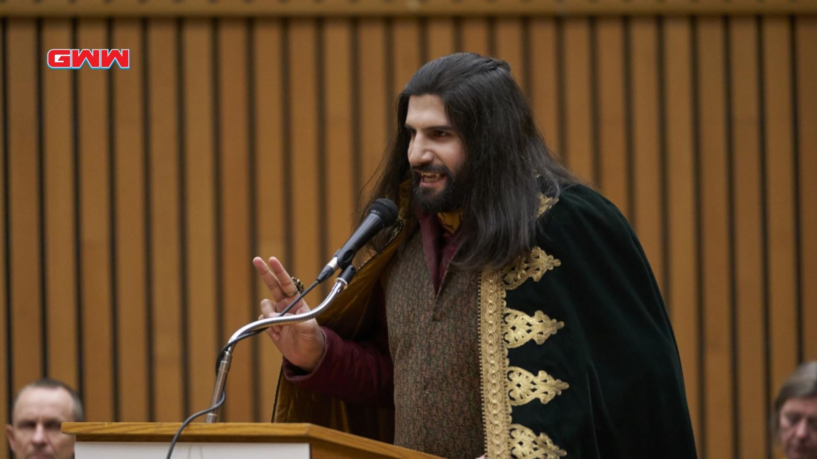 Nandor with long hair speaking at a podium in a formal setting.