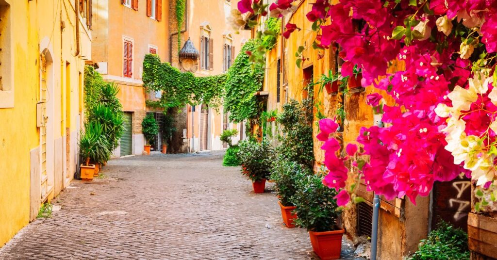 An Italian street full of orange and yellow concrete buildings draped with green ivy and pink and white flowers.