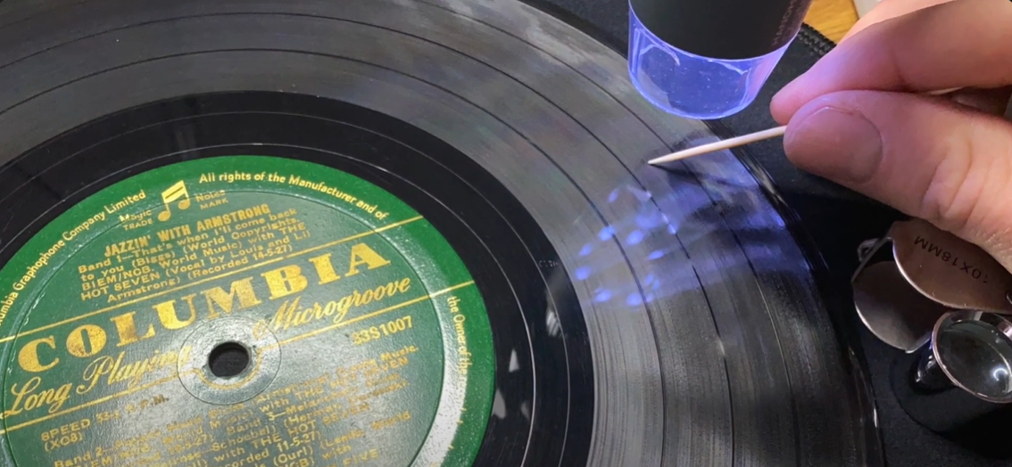 A person cleaning a vinyl record's grooves with a toothpick and magnifying tool.