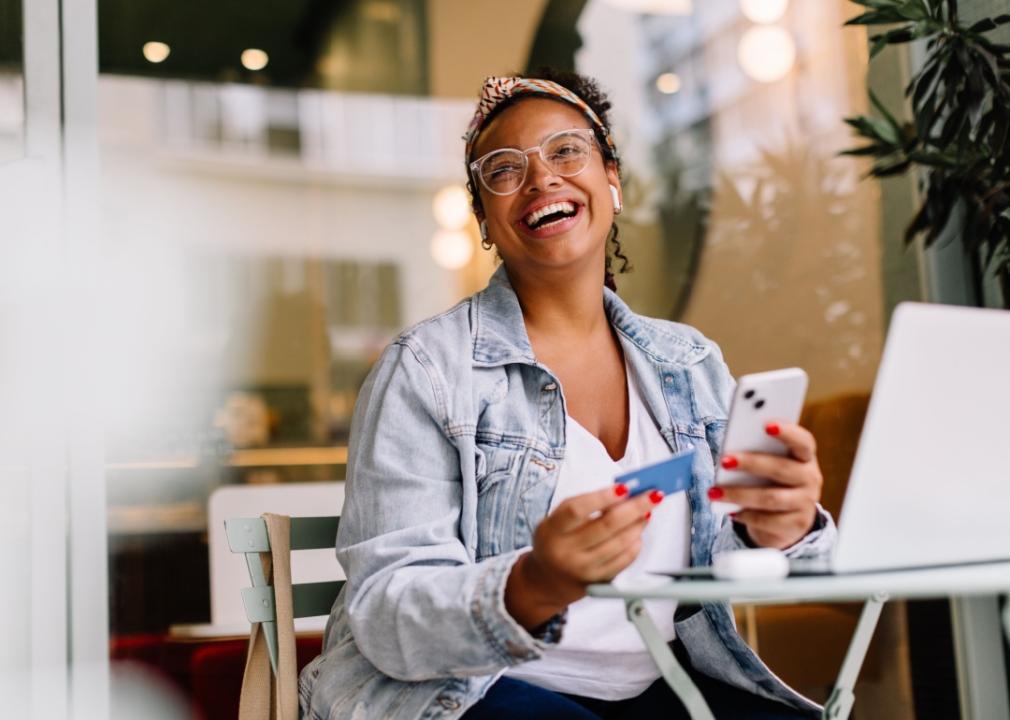 A woman with a bright smile sits at a table, holding a credit card in one hand and a smartphone in the other.