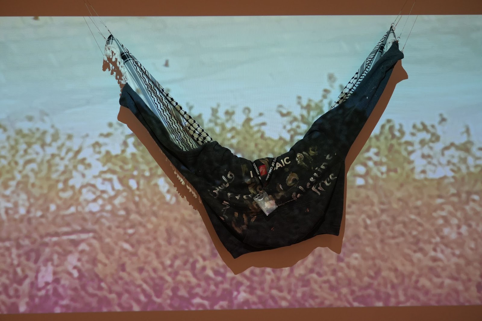 Image: A black shirt with white text hangs in front of a projected image of trees. Hung alongside the shirt is a black and white keffiyeh. An SAIC lanyard is draped over the garments. Photograph by Curators Under Censorship.