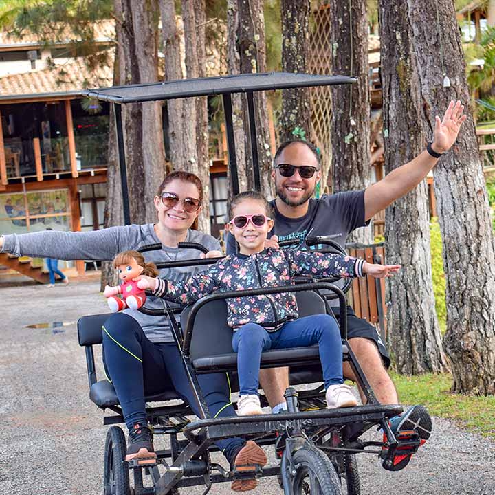 Família sorridente pedalando em um carrinho de pedal em meio à natureza com árvores ao fundo, aproveitando um momento de lazer ao ar livre.