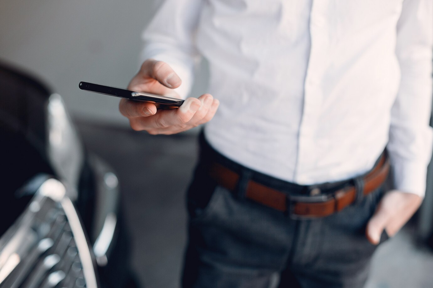 a person wearing a white shirt and dark trousers with a brown belt, holding a smartphone in one hand while the other hand is tucked into their pocket