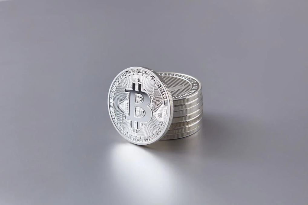 Light grey background with a stack of seven silver cryptocurrencies, topped with a silver Bitcoin coin, showcasing digital currency trends.