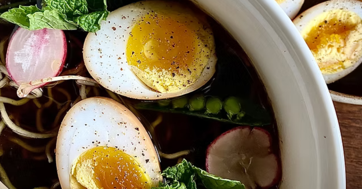 (Nahaufnahme von Ramen mit halbierten weichgekochten Eiern, Radieschenscheiben, Zuckerschoten, Minzblättern und gemahlenem schwarzen Pfeffer in einer kräftigen Brühe.)