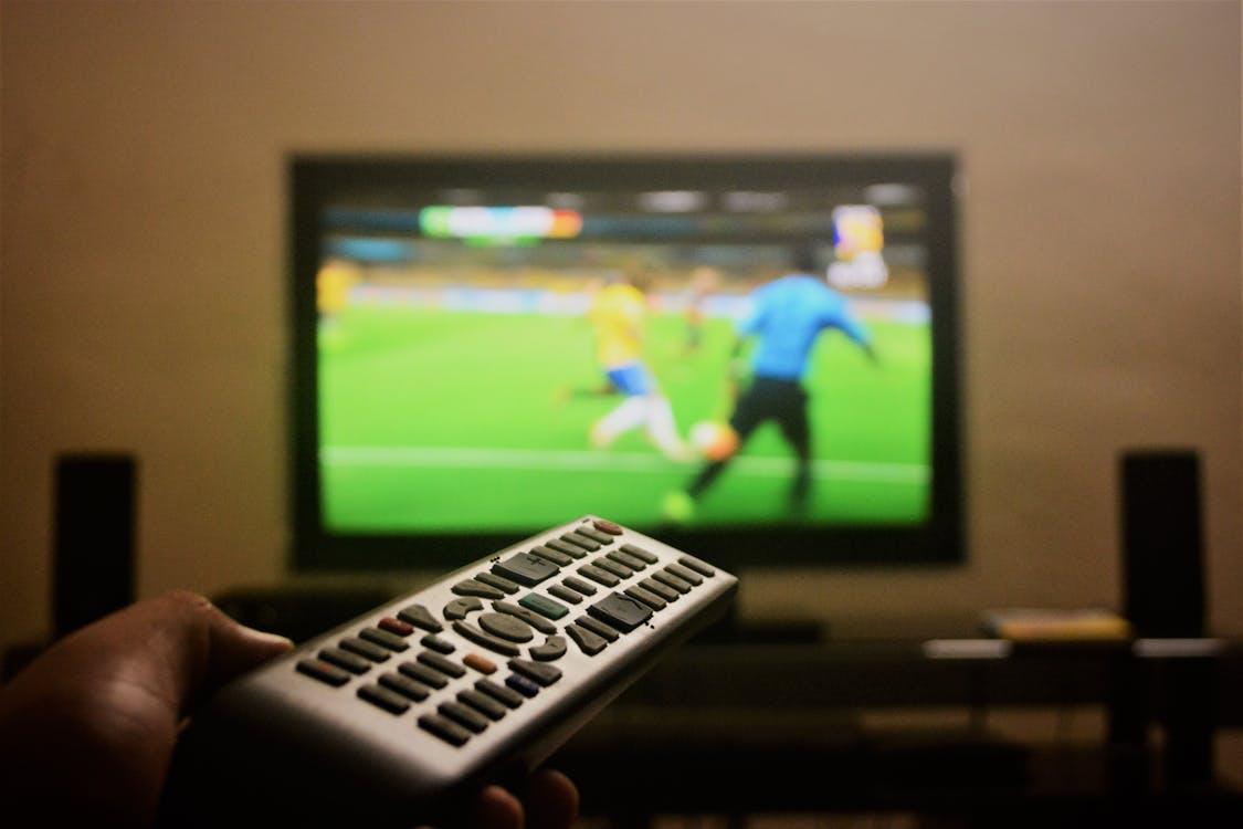 Free Close-up of a hand holding a remote control in front of a TV showing a football match. Stock Photo