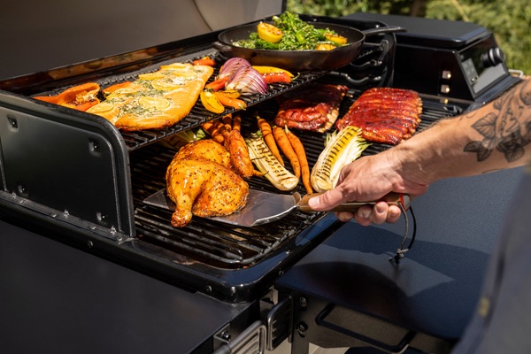 Man with metal meat flipper, flipping a piece of chicken on a Traeger grill.