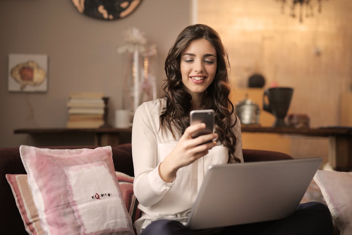Gratis Mujer Sentada En El Sofá Mientras Mira El Teléfono Con El Portátil En El Regazo Foto de stock