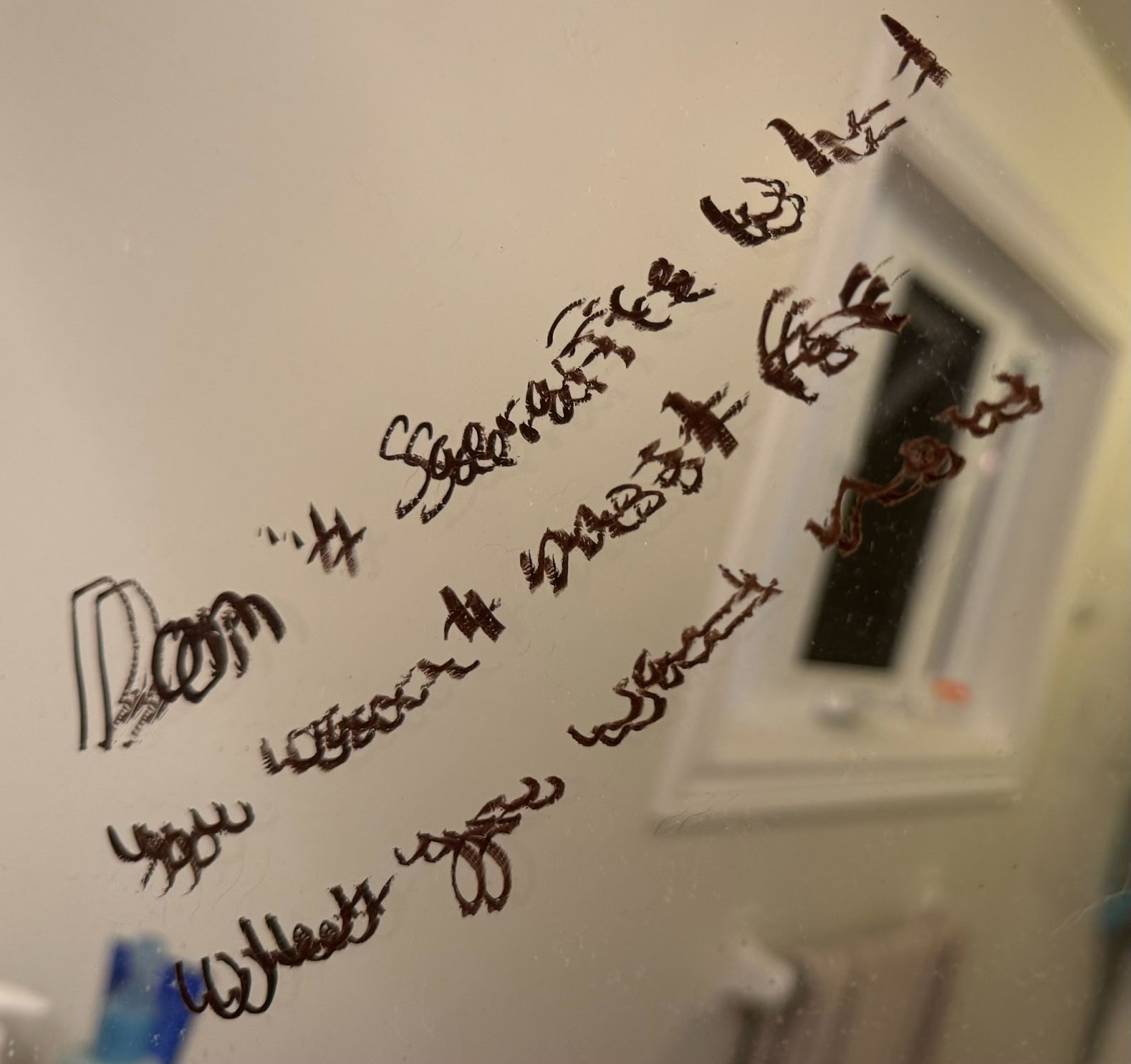 A close-up image of a bathroom mirror with a message written in dark brown eyeliner. The text appears to be handwritten in cursive, but it is smudged and difficult to read clearly. The reflection in the mirror shows a blurred window and part of the bathroom interior, including a towel rack and some indistinct objects in the foreground. The lighting is soft, giving the scene a warm, dim glow. The text in the mirror seems to reference "sacrifice" and "must be," but the rest of the writing is unclear due to the smudging.