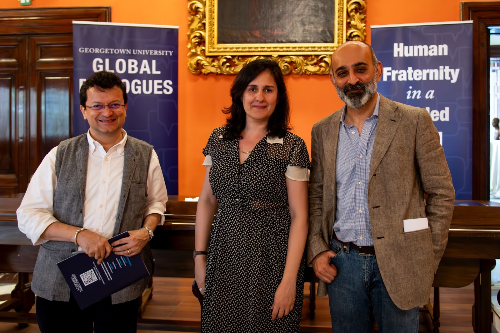 Kamila stands between fellow authors  Ranjit Hoskote and Mohsin Hamid in front of conference branding and a gold framed painting. They are all smiling and standing casually.